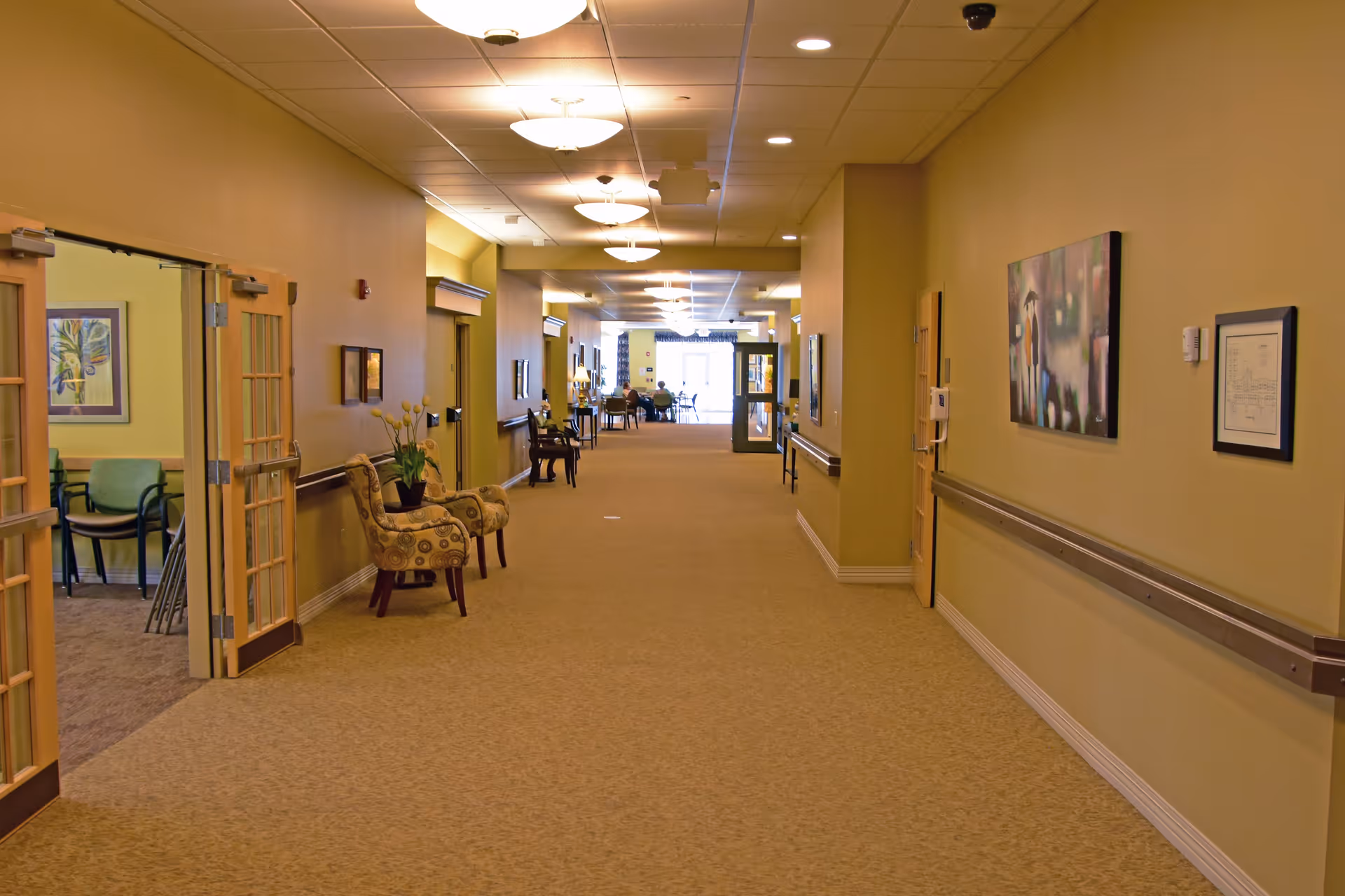 A long, well-lit hallway in an assisted living facility with beige walls and carpet. There are chairs and small tables with plants along the walls, framed artwork, and open double doors leading to a room with additional chairs. At the far end, there are more chairs and a glass door letting in natural light.