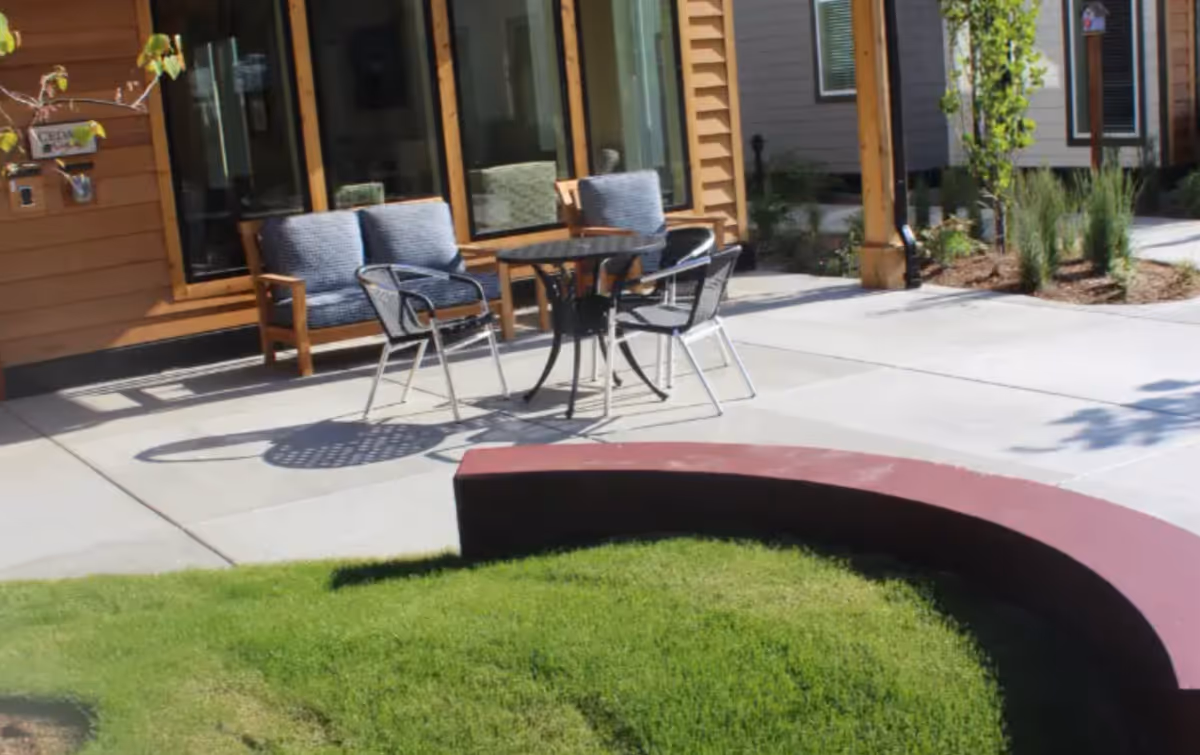 Outdoor patio with a round table, several chairs, a wooden bench, and a curved planter edging a grassy area in front of a building.