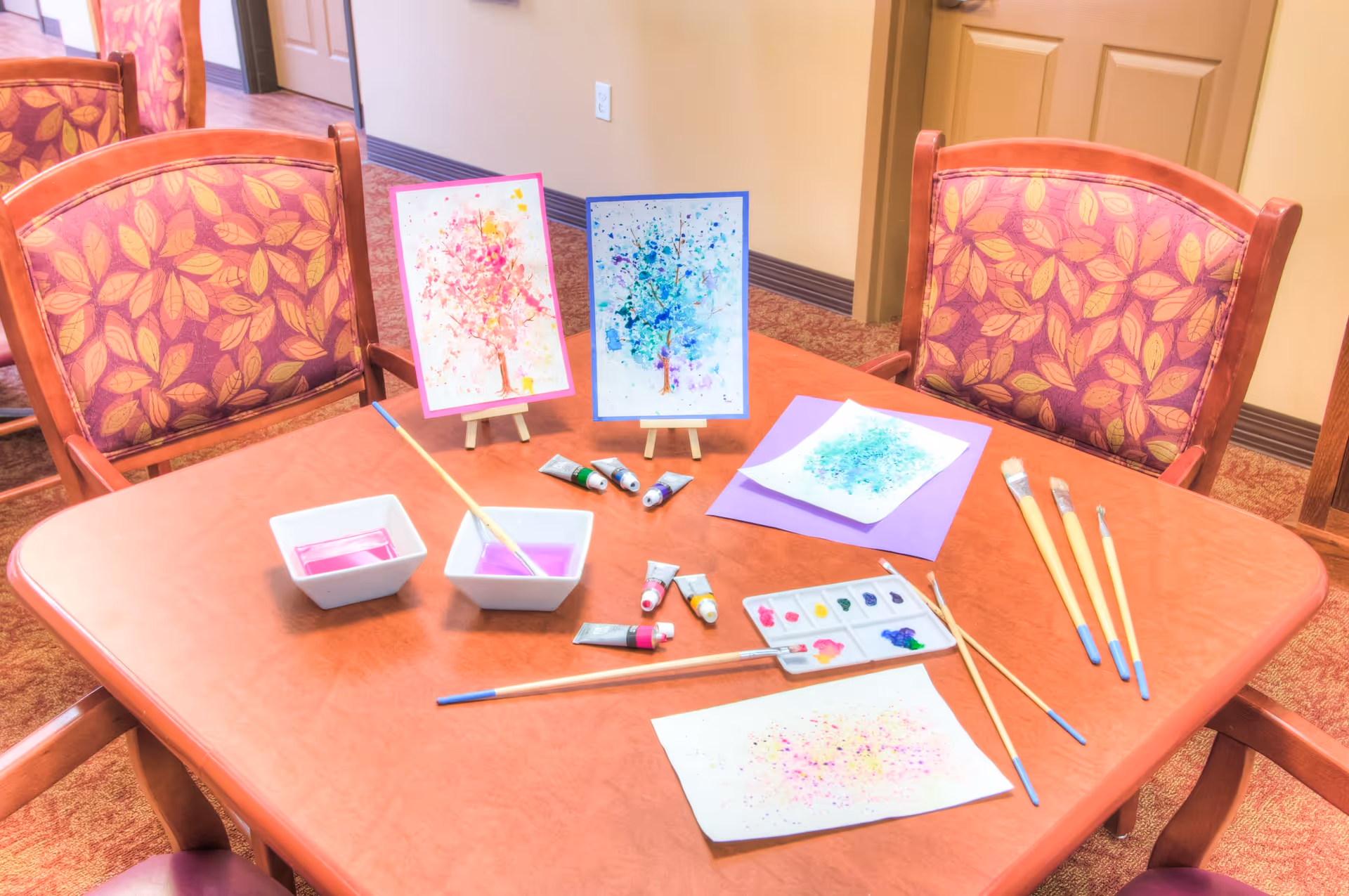 A wooden table in a room with four chairs featuring leaf-patterned upholstery. On the table are watercolor paintings of trees, paintbrushes, paint tubes, two bowls with purple water, and a paint palette with various colors. The setting appears to be a creative activity area within a senior living facility.