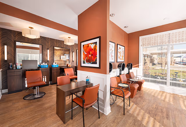 Bright, well-lit salon area with orange styling chairs, a manicure table, seated hair dryers, wood floors and framed artwork beside a large window.