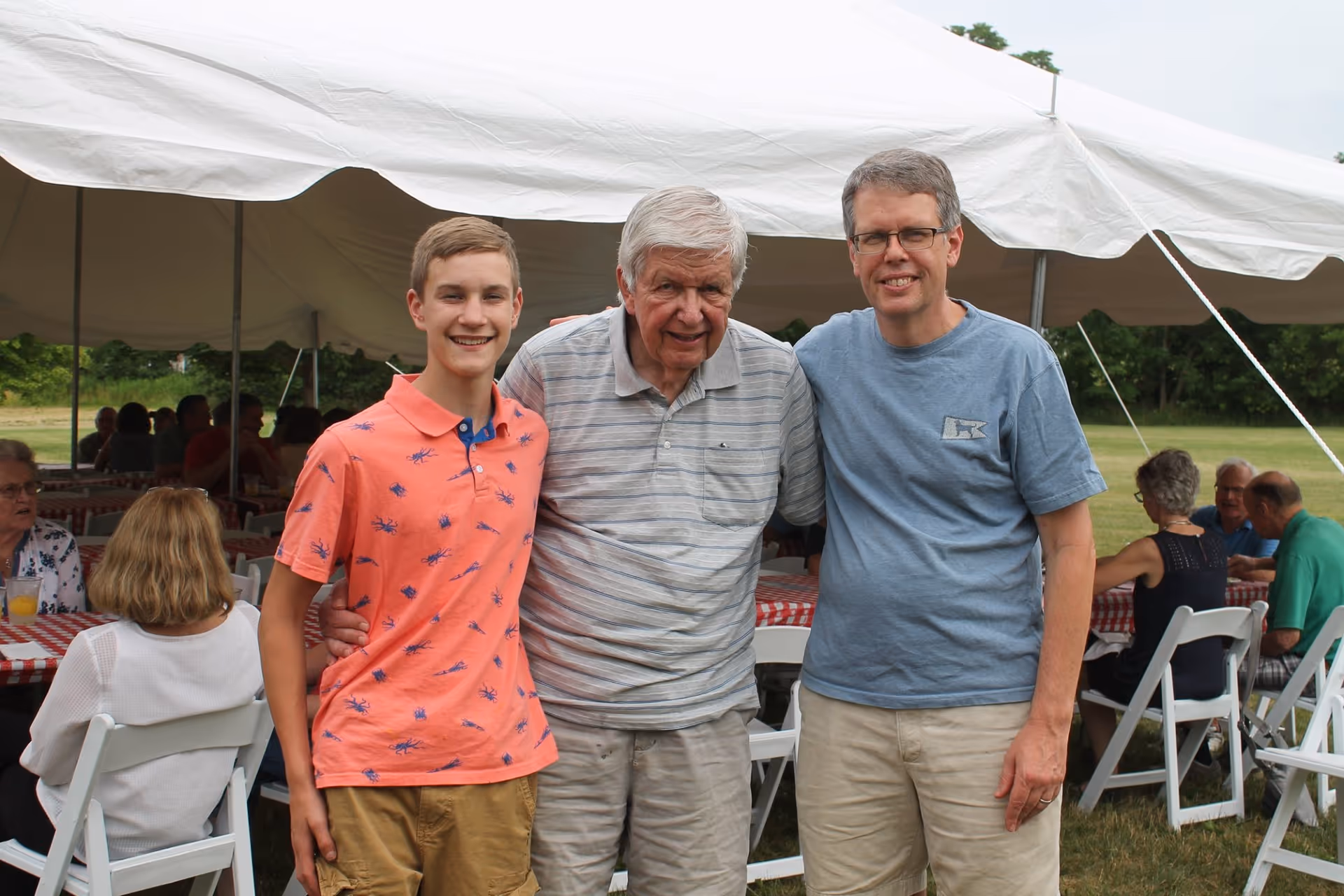 Three males of different generations standing together and smiling under a large white outdoor tent with people seated at tables covered with red and white checkered tablecloths in the background.