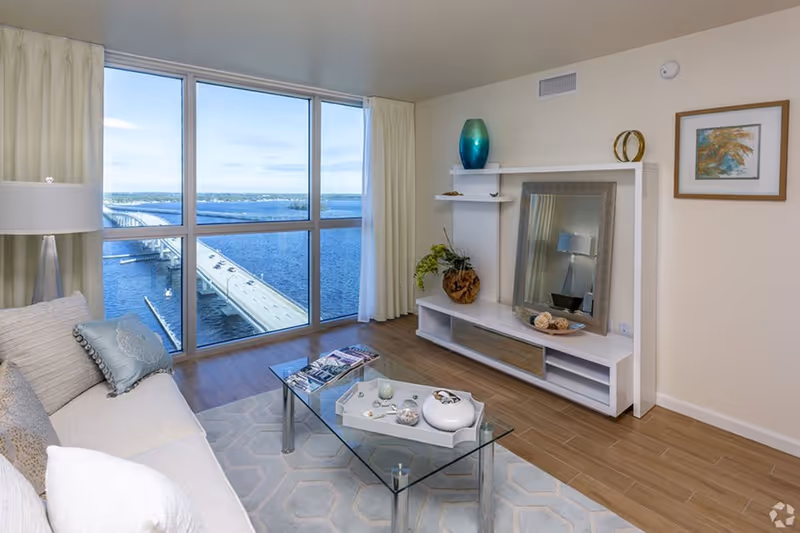 A bright living room with a large window offering a view of a bridge over water. The room features a beige sofa with decorative pillows, a glass coffee table with decorative items, a white entertainment unit with a large mirror and decorative vases, a floor lamp, and a framed artwork on the wall.