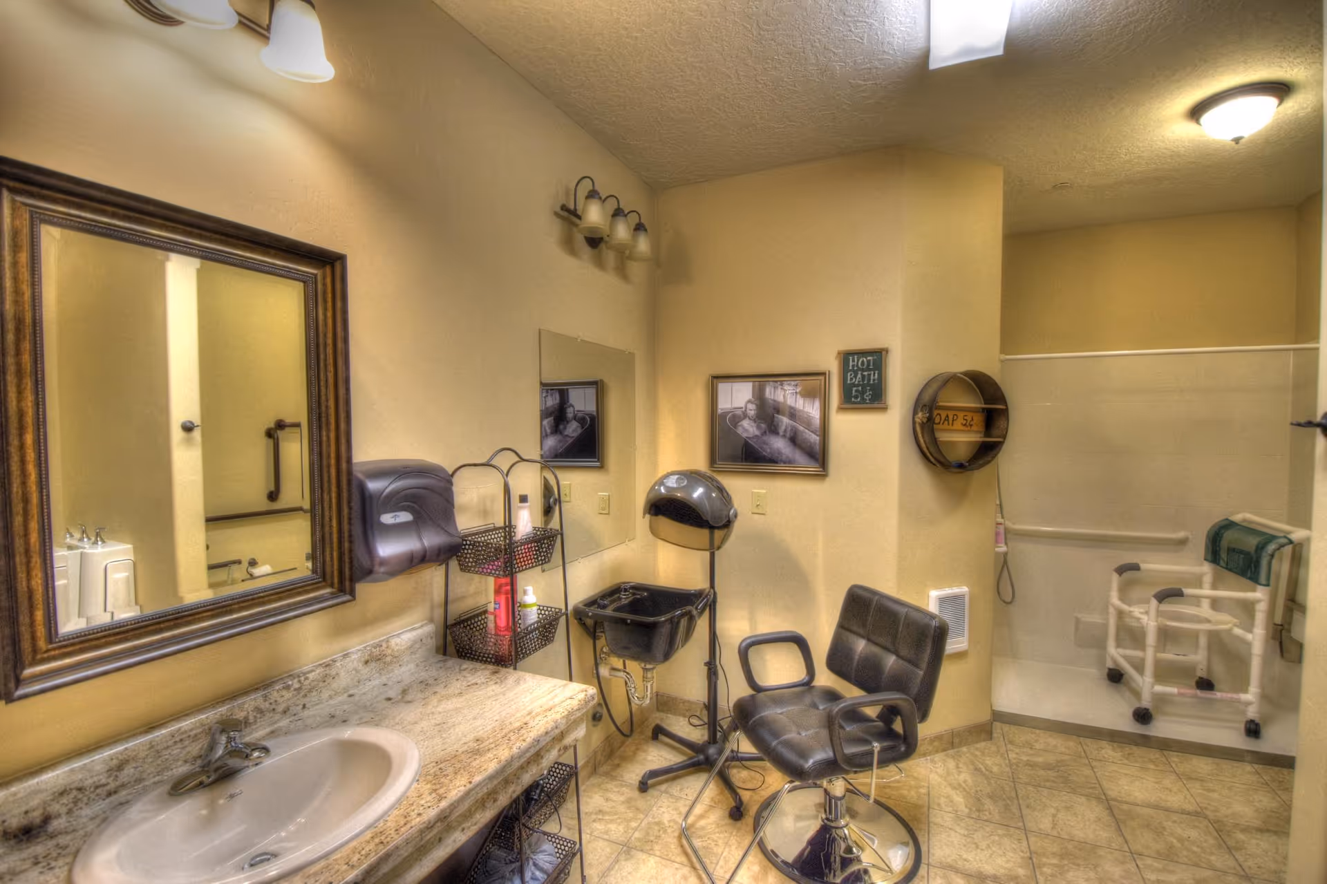 Interior view of a bathroom area in a senior living facility featuring a sink with a large mirror above it, a black salon chair, a hair dryer hood, and a shower area with a white shower chair and grab bars. The walls are beige, and there are framed pictures and small signs on the walls.