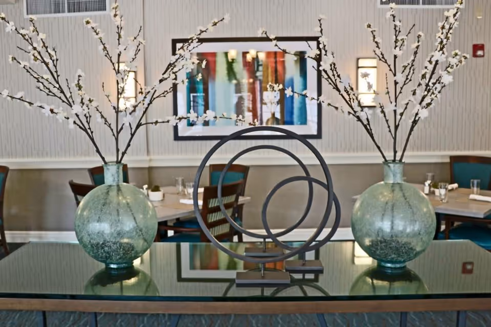 A decorative arrangement on a glass table featuring two large green glass vases with white flowering branches and a modern metal sculpture with three concentric circles. In the background, there are dining tables with chairs, a colorful abstract painting on the wall, and wall-mounted lights.