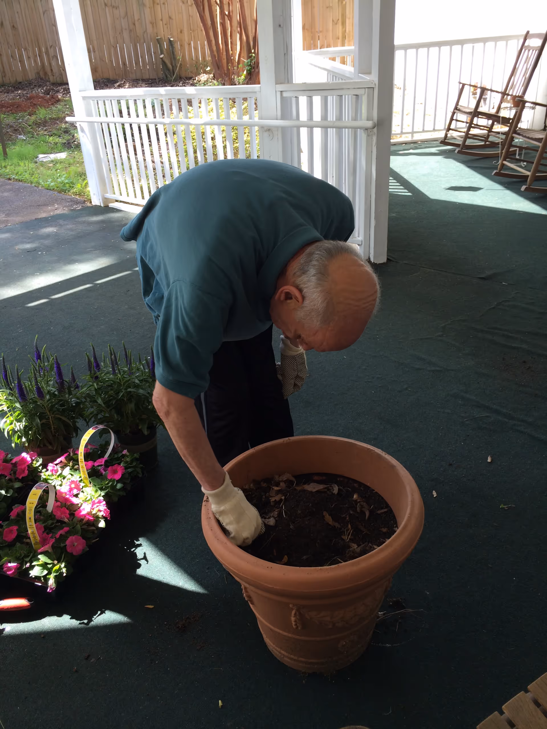An elderly man wearing gardening gloves is bent over, working with soil in a large terracotta planter on a covered porch. Nearby are several pots of flowers ready to be planted. The porch has a green carpet and white railing, with rocking chairs in the background.