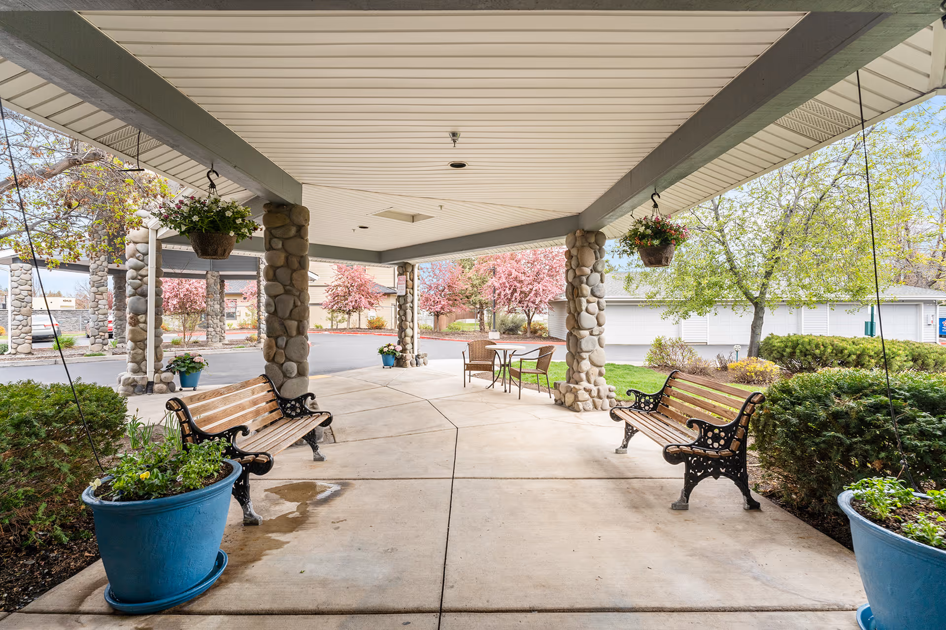 Covered outdoor seating area with two wooden benches with black metal armrests and legs, hanging flower pots, large blue plant pots with greenery, stone pillars supporting the roof, and a small table with two chairs in the background. Surrounding the area are green bushes, trees with pink blossoms, and a paved driveway.