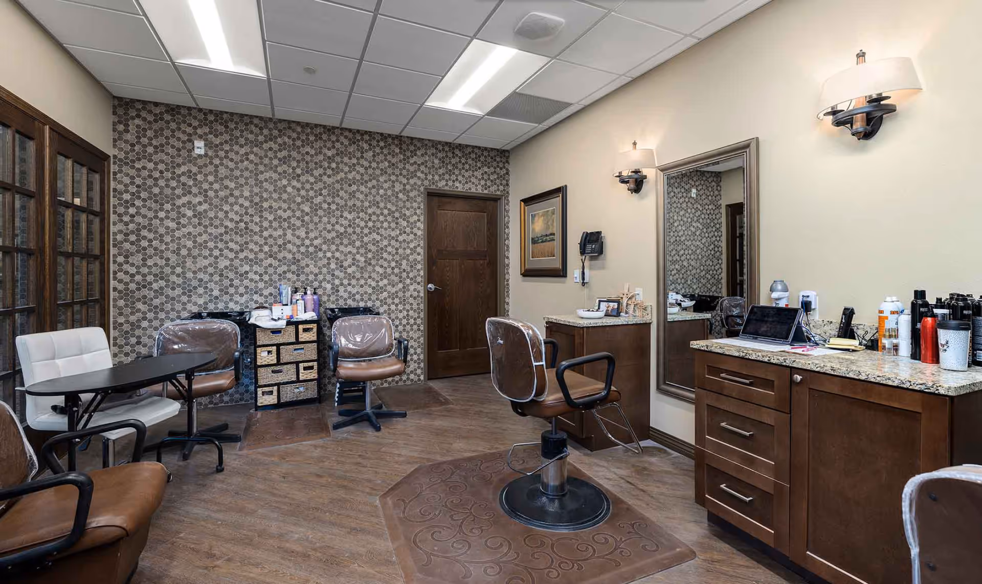 Interior view of a salon area in a senior living facility with brown leather styling chairs, a large mirror, wooden cabinets with granite countertops, hair care products, and a small table with chairs. The room has a patterned accent wall, wood flooring, and ceiling lights.