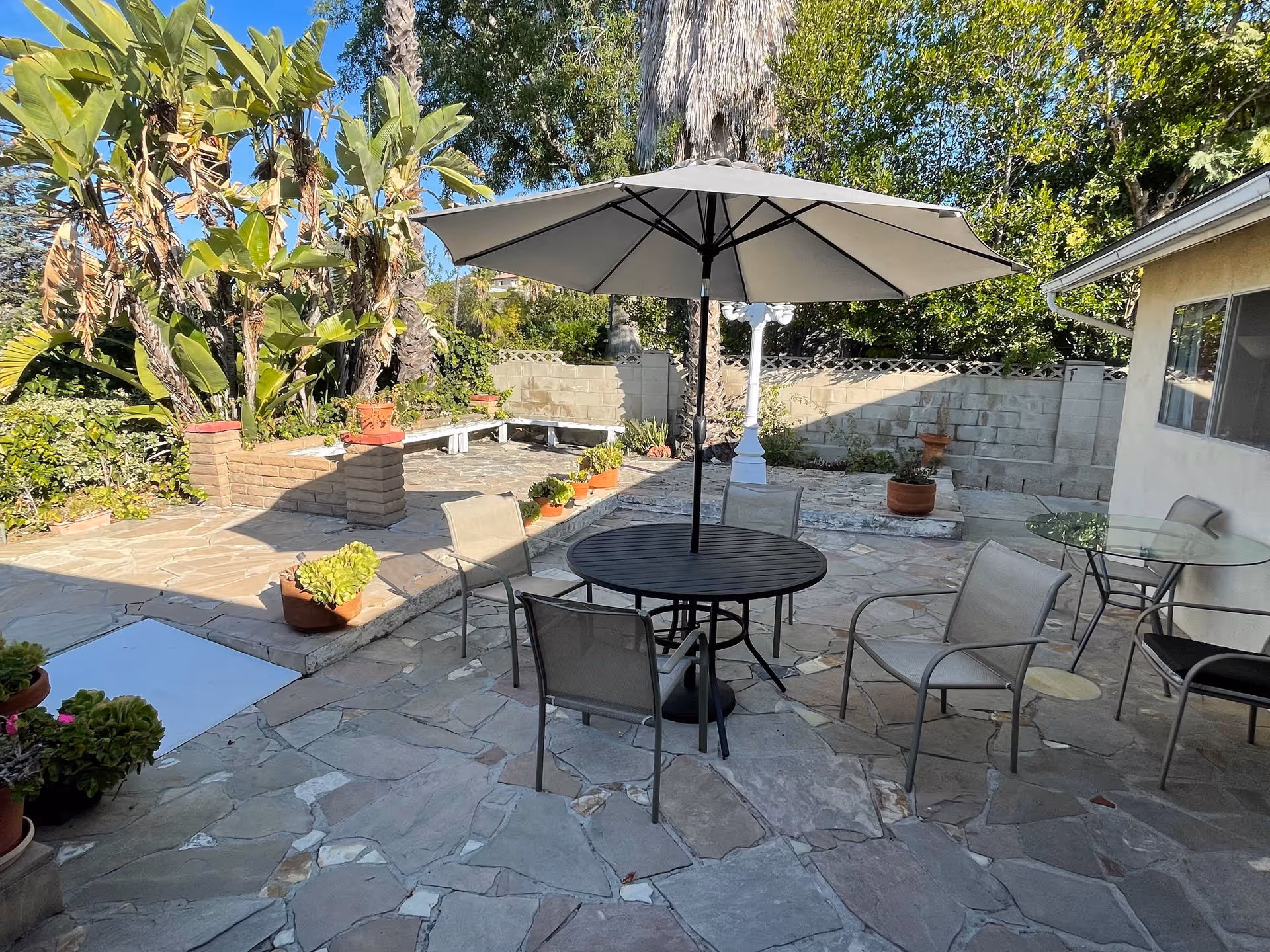 Outdoor patio area with stone flooring, a round black table with a large gray umbrella, and several chairs around it. There are potted plants placed on the ground and on raised brick planters. A glass table with chairs is positioned near the building wall, and trees and greenery surround the area.