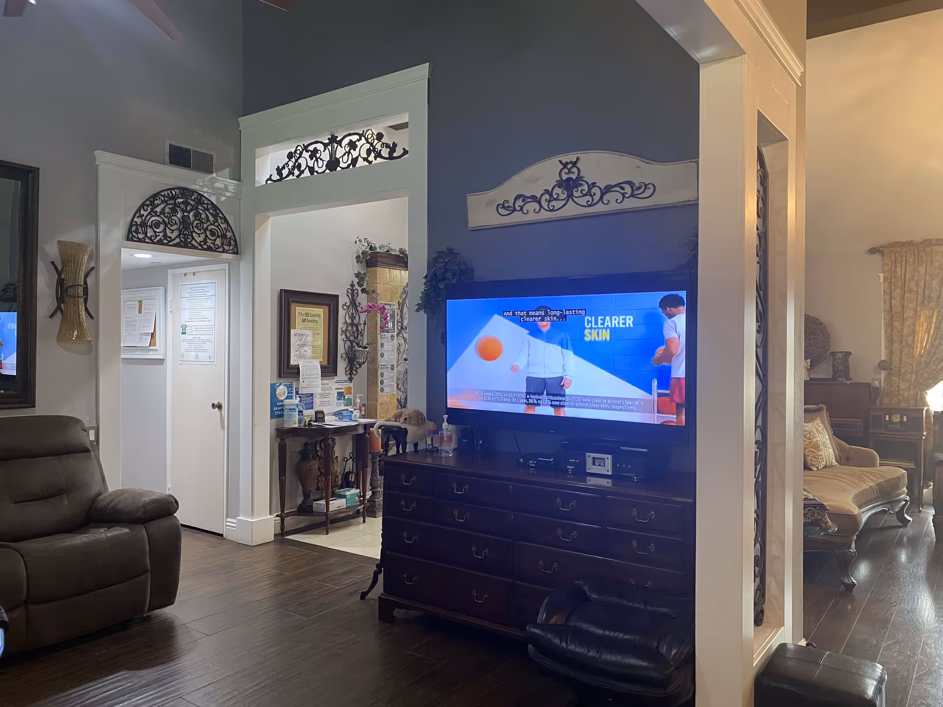Interior view of a living room area in a senior living facility with a large flat-screen TV mounted on a dark wooden dresser. The TV is displaying a commercial about clearer skin. To the left, there is a brown recliner chair and a small table with hand sanitizer and notices on the wall. The room has dark wooden flooring and decorative wrought iron accents above doorways and on the walls. In the background, there is a seating area with a beige upholstered sofa and curtains.