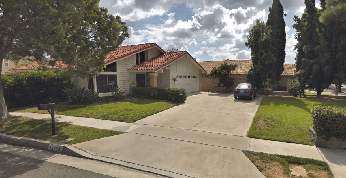 A suburban house with a red-tiled roof, white exterior walls, a two-car garage, and a driveway with a parked car. The front yard has green grass, bushes, and trees under a partly cloudy sky.
