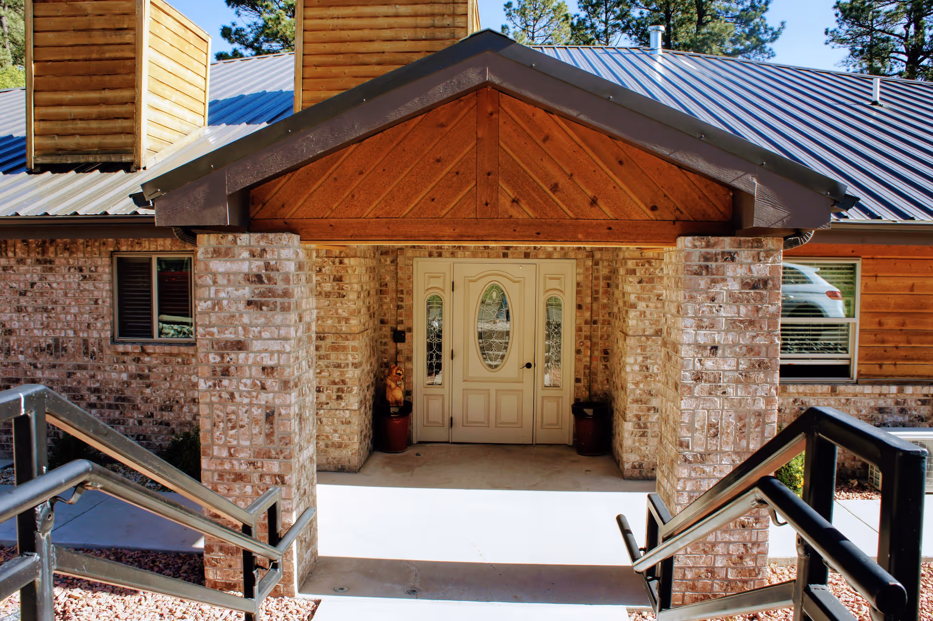 Covered front entrance of a brick building with double decorative doors, brick columns, and metal handrails leading up to it.