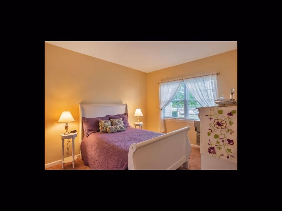 Cozy bedroom with a white sleigh bed dressed in purple bedding, two bedside tables with lamps, a window with sheer curtains, and a decorative dresser.
