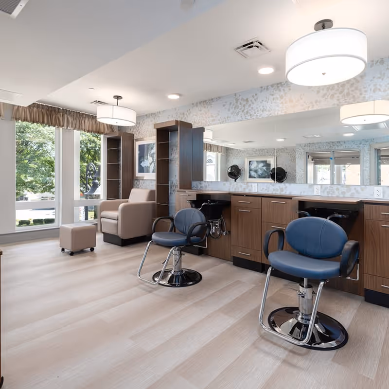 Bright and modern salon area with two blue salon chairs in front of a long mirror and wooden cabinetry. A beige armchair and matching ottoman are positioned near large windows with a view of green trees outside. The room has light wood flooring and ceiling lights.