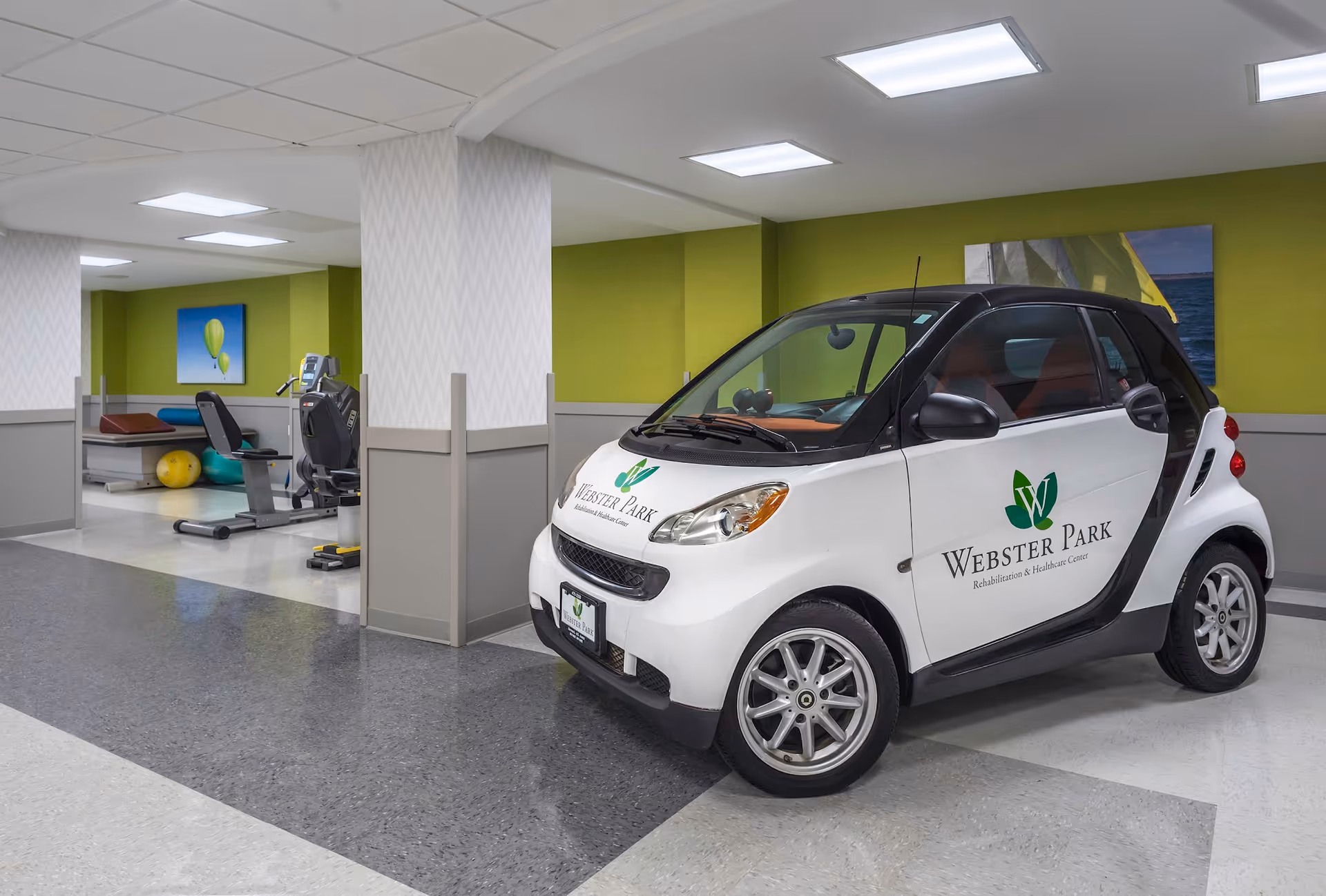 Small white and black Smart car with Webster Park Rehabilitation & Healthcare Center logo parked inside a facility hallway near exercise equipment and therapy balls.