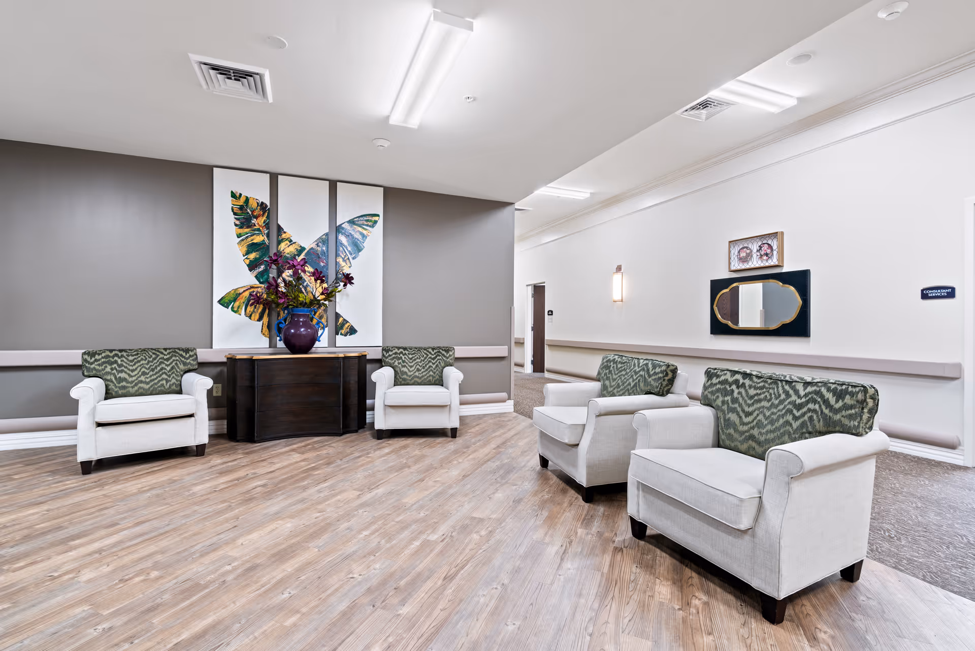 A bright and clean common area with four white armchairs featuring green patterned cushions arranged around a dark wooden console table with a large vase of flowers. Behind the table is a three-panel wall art piece depicting a colorful butterfly. The room has light wood flooring, gray and white walls, and ceiling lights.