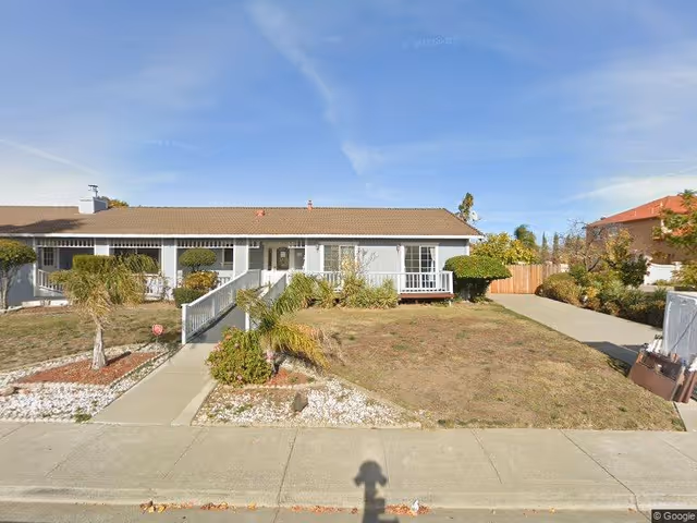 Single-story light-blue farmhouse-style building with a front ramp and porch, dry lawn, and driveway under a clear blue sky.