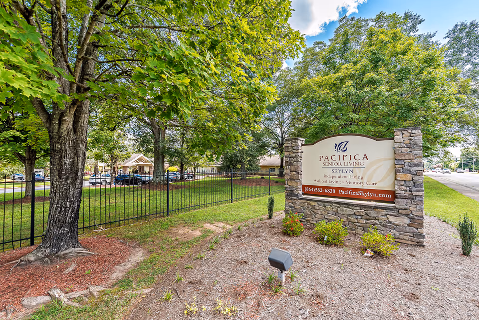 Outdoor view of a senior living facility entrance with a stone sign that reads 'Pacifica Senior Living Skylyn Independent Living Assisted Living Memory Care' surrounded by trees, grass, and a black metal fence. Cars and a building are visible in the background.