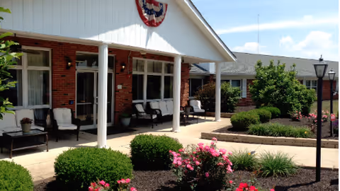 Front entrance of a brick senior living facility with a covered porch, outdoor seating, and landscaped flowerbeds.
