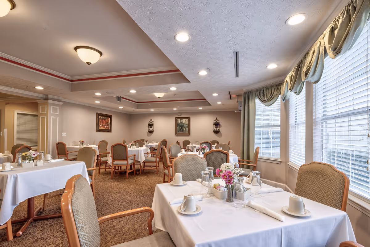 Well-lit dining room with multiple tables set with white tablecloths, chairs, and large windows with drapes.
