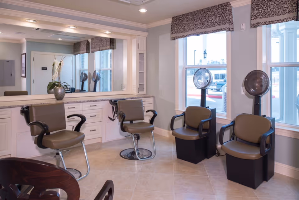 Interior view of a salon area in a senior living facility with two salon chairs in front of a large mirror and two hair drying chairs positioned near windows with patterned valances.