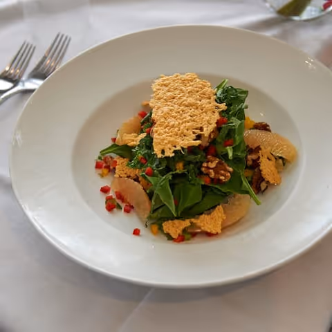 Plated salad of greens with grapefruit segments, walnuts, diced red pepper and crispy cheese shards on a white plate set on a white tablecloth next to silverware.