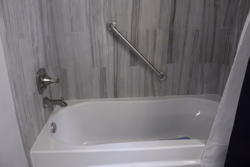 White bathtub with gray wood-look tile surround, chrome faucet, and a diagonal grab bar on the wall.