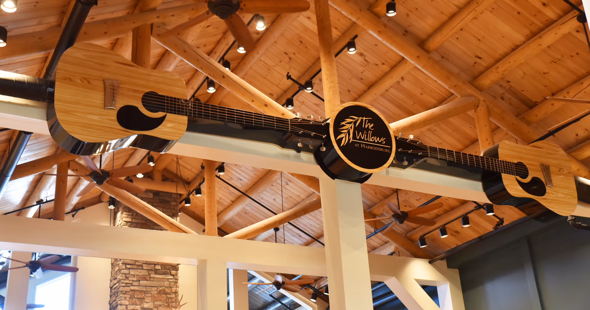 Interior view of a wooden-beamed common area ceiling decorated with a large guitar sign reading 'The Willows at Harrodsburg'.