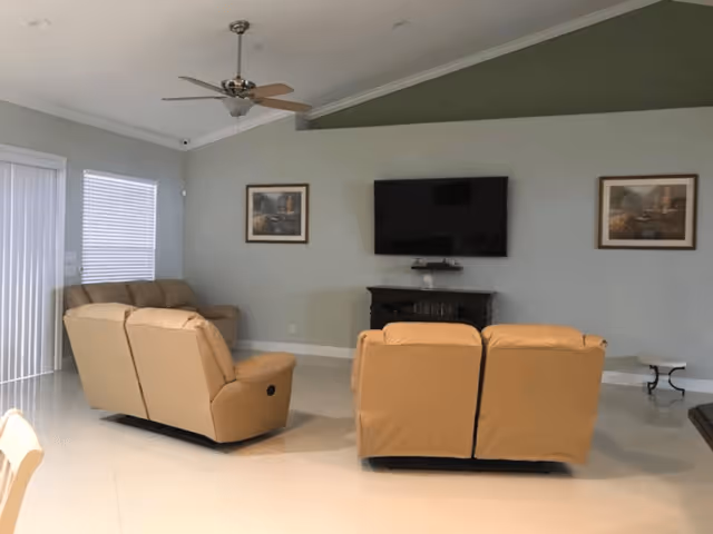 A living room with beige leather recliner sofas arranged facing a wall-mounted flat-screen TV. The room has light-colored tiled flooring, a ceiling fan with light, two framed pictures on the wall, and a small table to the right. There are windows with blinds and a sliding glass door on the left side.
