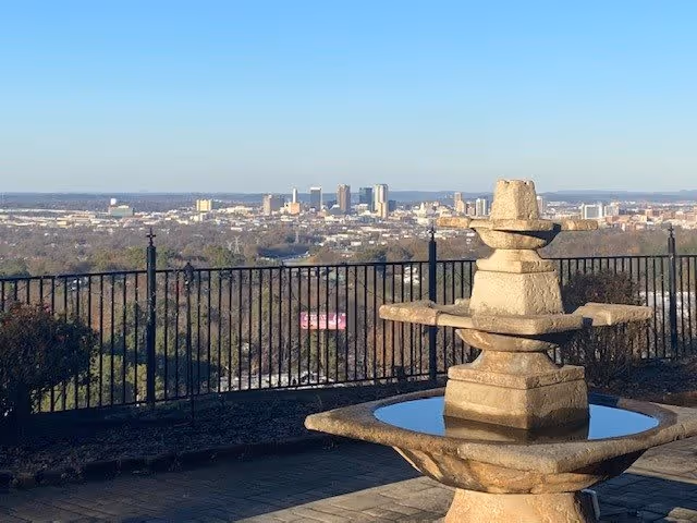 Stone water fountain in the foreground with a black metal fence behind it, overlooking a city skyline in the distance under a clear sky.