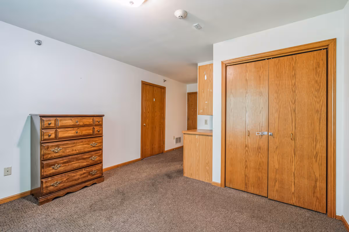 Empty bedroom with a wooden dresser, carpeted floor, and double-door closet.