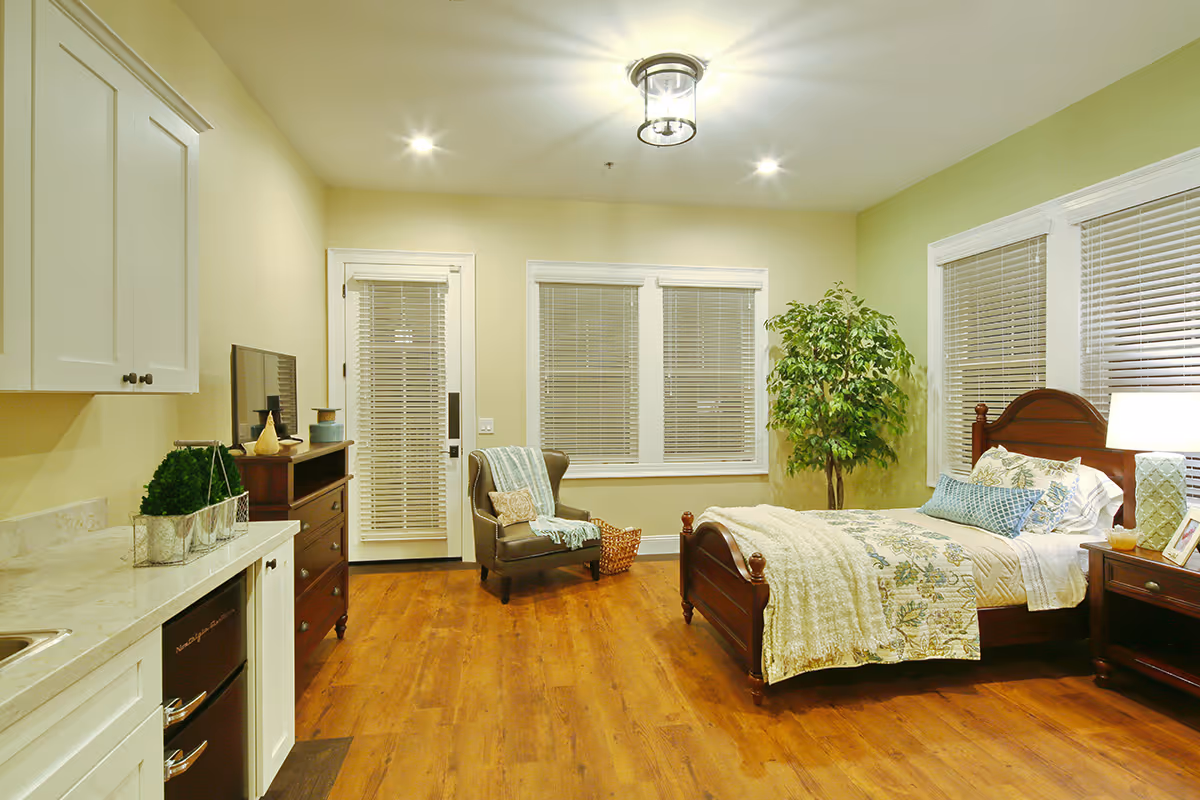 A well-lit bedroom in an assisted living facility with a wooden bed, nightstand, armchair, small kitchenette area, and a potted plant.