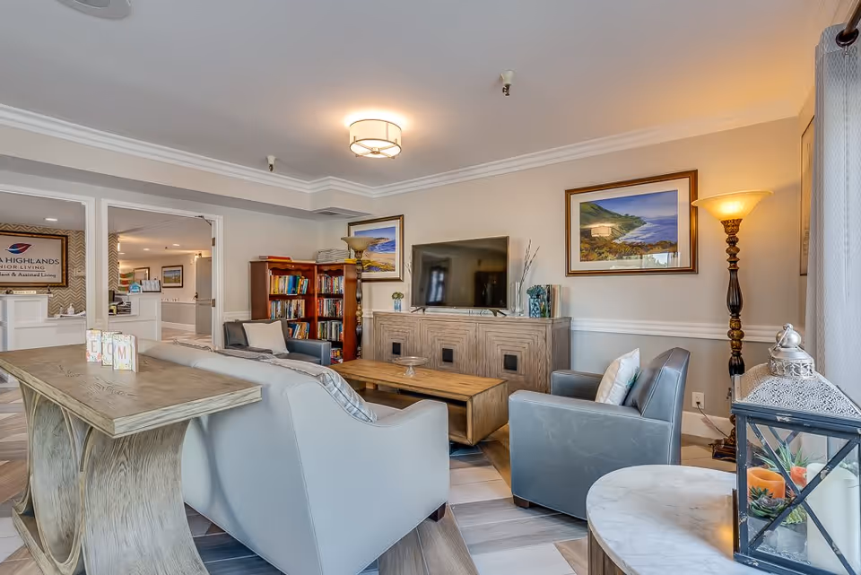 A cozy senior living facility common area with a light gray sofa, two gray armchairs, a wooden coffee table, a TV on a wooden cabinet, a bookshelf filled with books, framed landscape paintings on the walls, and a tall floor lamp. The room has light-colored walls and a ceiling light fixture. In the background, a reception area with a sign that reads 'Serra Highlands Senior Living' is visible.