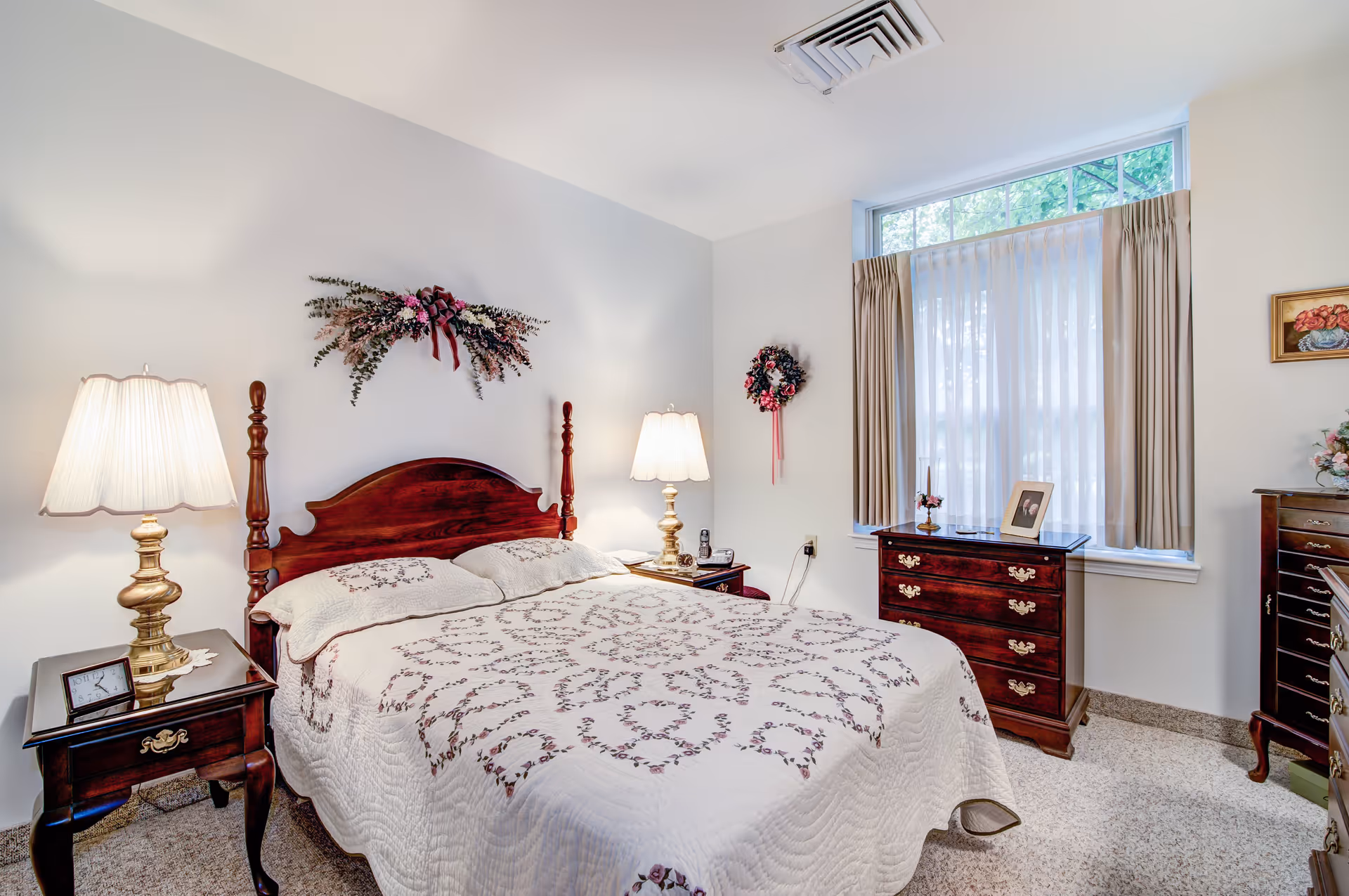 A cozy bedroom with a wooden bed frame and a white quilted bedspread featuring a floral pattern. Two bedside tables with brass lamps flank the bed. The room has a large window with beige curtains and sheer white drapes, allowing natural light to enter. Floral wreaths and decorations adorn the walls, and there are wooden dressers with decorative items and framed photos.