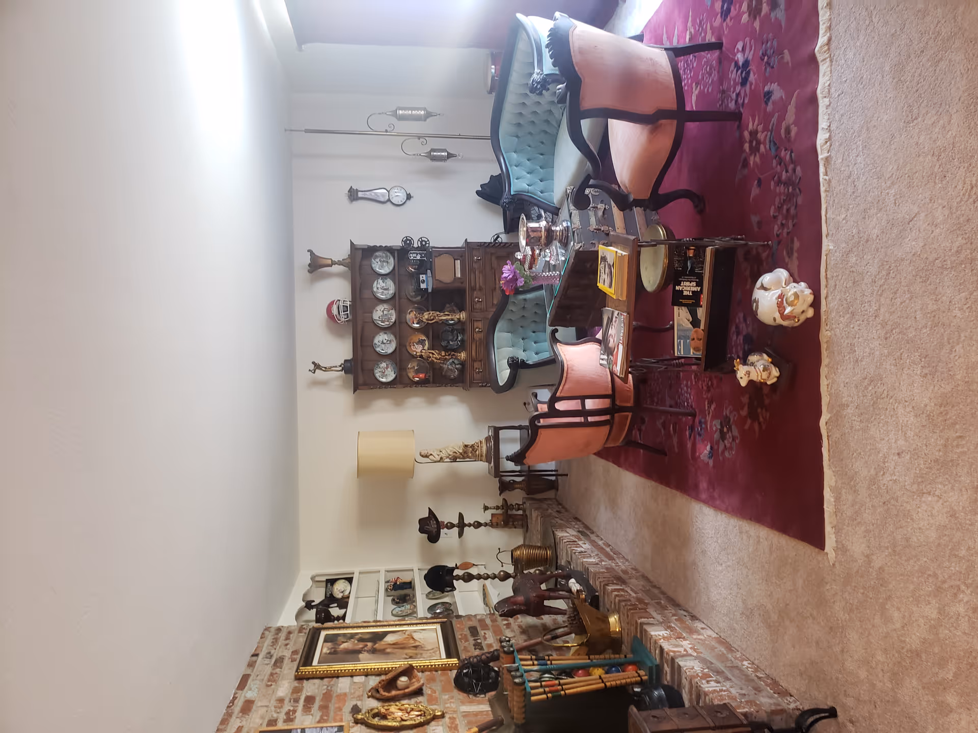 A cozy living room with vintage furniture including two light blue tufted sofas and two pink upholstered chairs arranged around a glass-topped coffee table on a red floral rug. The room features a brick fireplace with various decorative items, a wooden cabinet displaying plates and figurines, a floor lamp, and wall-mounted clocks and lanterns. The room is softly lit by natural light coming through a window with curtains on the right.