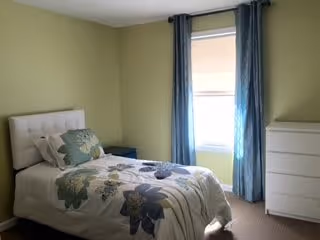 A small bedroom with a single bed covered in a floral patterned comforter and pillows. The room has light green walls, a window with blue curtains, and a white dresser to the right of the bed.