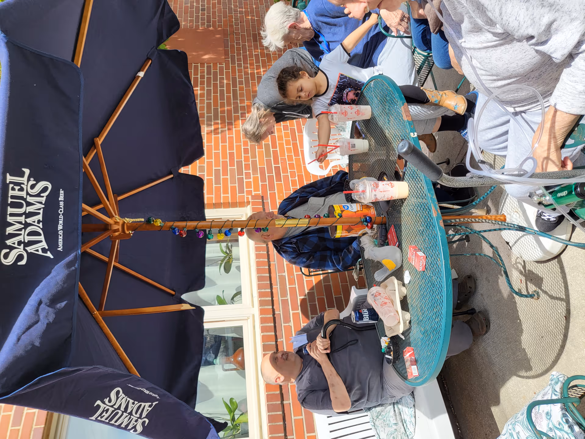 People seated around a round metal patio table under a Samuel Adams umbrella on a brick exterior patio.