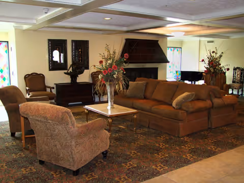 A cozy living room area with a large brown sectional sofa, two upholstered armchairs, a wooden coffee table with a vase of flowers, a fireplace with a dark wooden mantle, and decorative framed mirrors on the wall. The room has a patterned carpet and soft lighting.