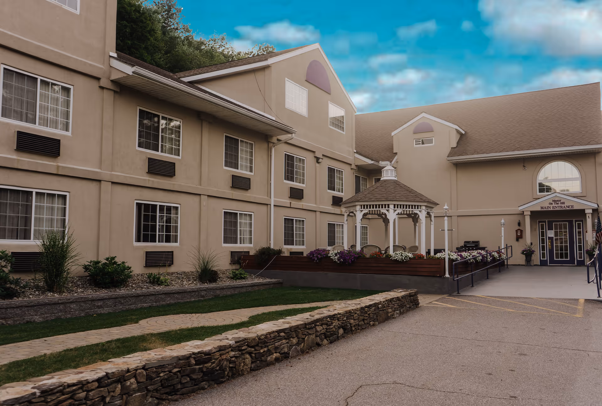 Exterior view of Manor On the Hill senior living facility showing a beige multi-story building with multiple windows, a small white gazebo surrounded by flowers, a stone wall, and a paved driveway leading to the main entrance under a blue sky with some clouds.
