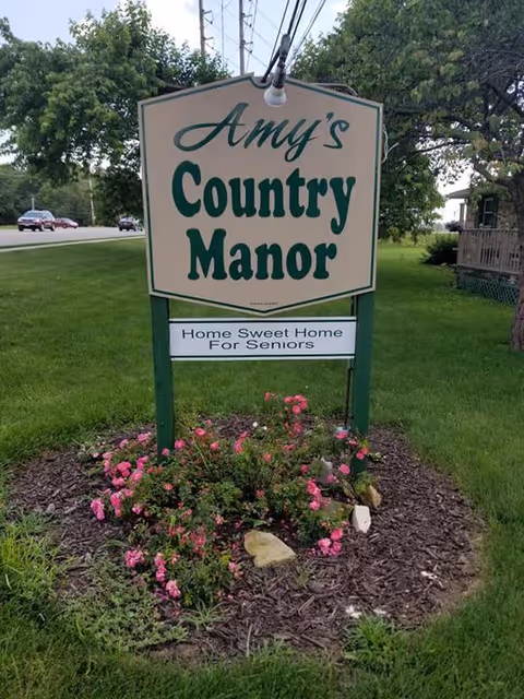 A freestanding yard sign reading "Amy's Country Manor - Home Sweet Home For Seniors" surrounded by grass and pink flowers.