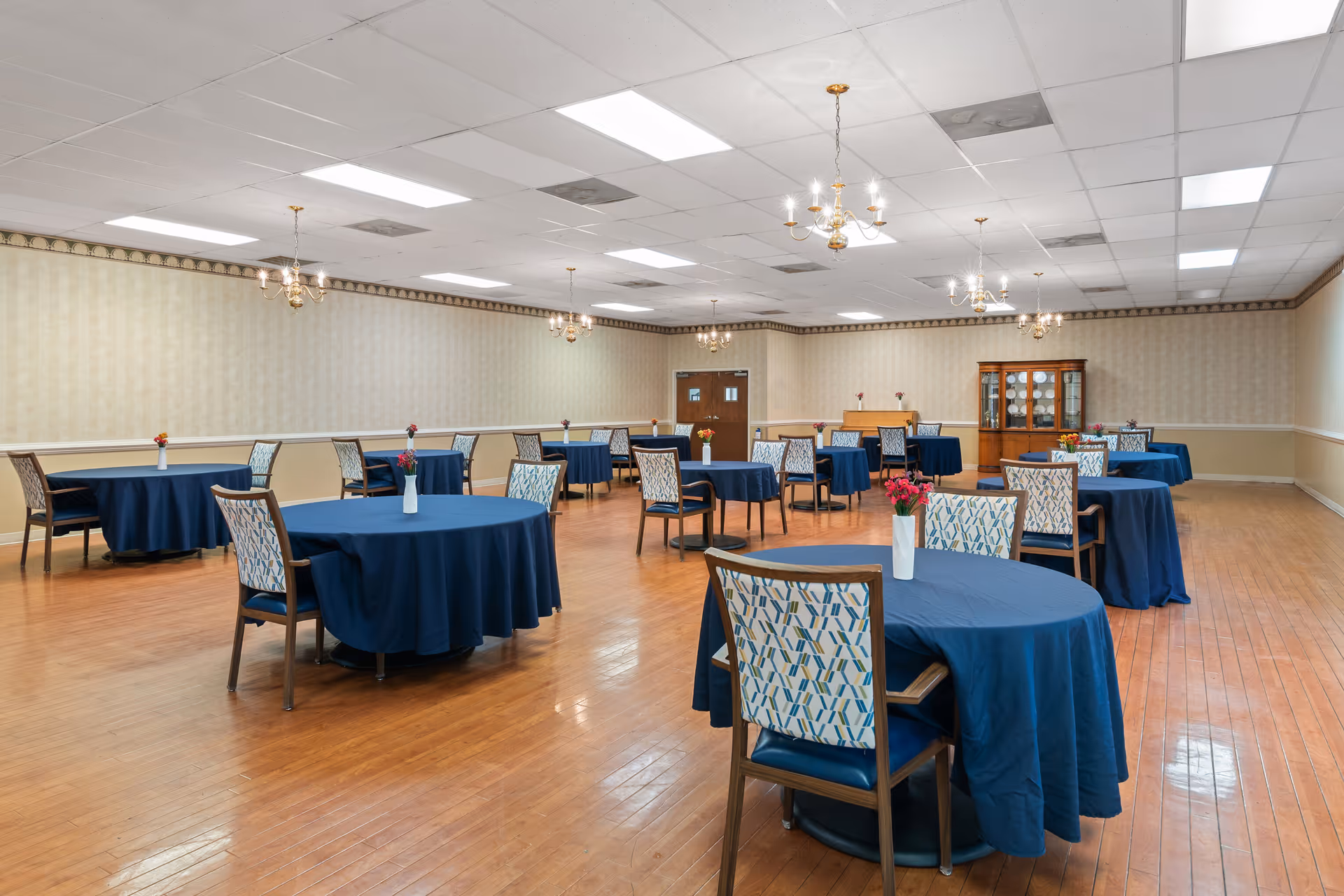 A spacious dining room with multiple round tables covered in dark blue tablecloths, each table surrounded by patterned chairs. Small vases with flowers are placed on each table. The room has wooden flooring, beige wallpaper with a decorative border, and several chandeliers hanging from the ceiling. A wooden cabinet with glass doors is visible at the far end of the room.
