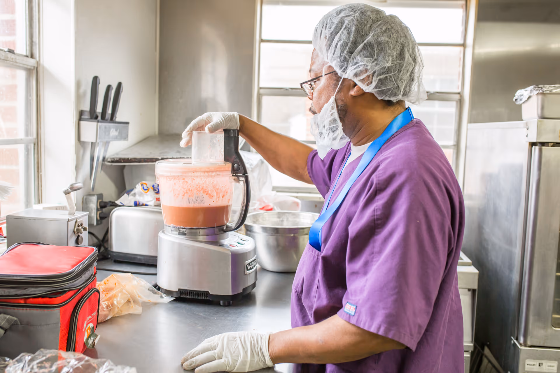 A person wearing a hairnet, gloves, and a purple uniform is operating a food processor with a pink mixture inside in a commercial kitchen setting.