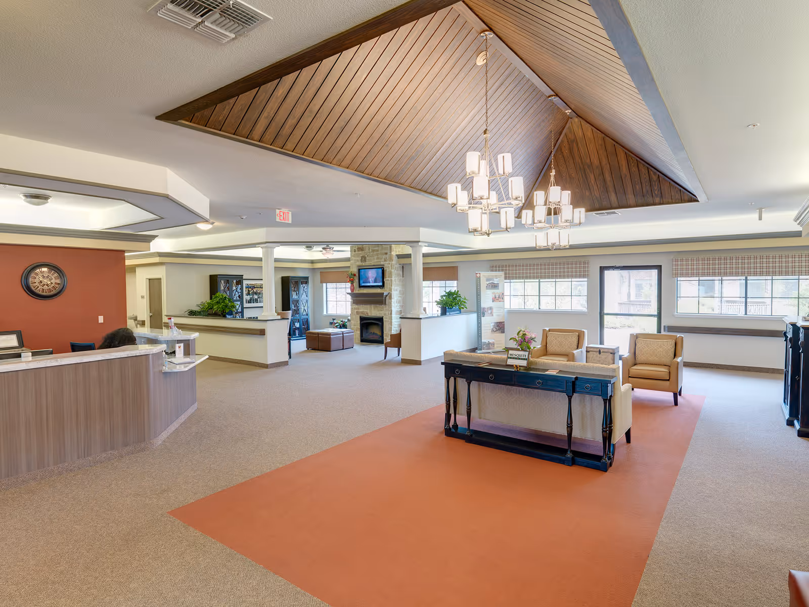 Spacious senior living facility lobby with a reception desk, seating area, fireplace, and chandelier under a wood-paneled ceiling.