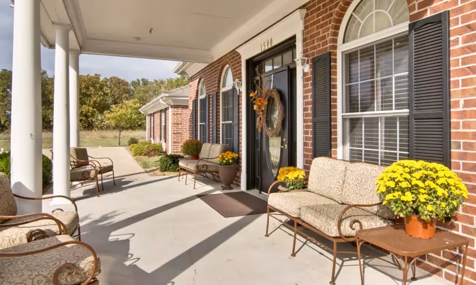 Covered brick front porch with columns, seating, potted yellow flowers, and a black entry door.