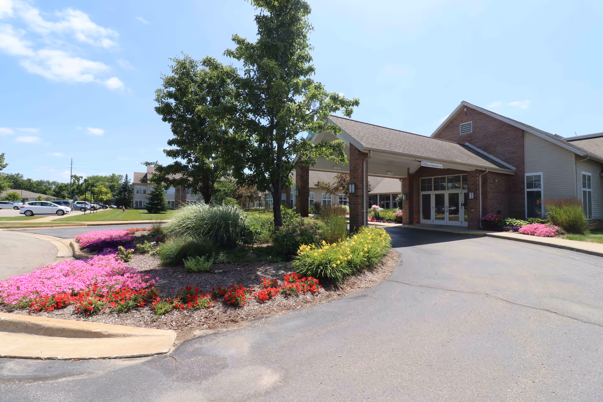 Entrance of Brecon Village senior living facility with a covered drop-off area, surrounded by landscaped flower beds with pink, red, and yellow flowers, green shrubs, and trees under a clear blue sky.