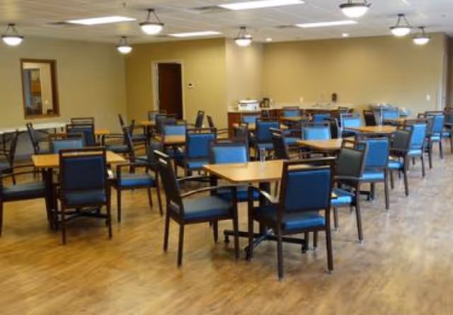 A spacious dining room with multiple wooden tables and blue cushioned chairs arranged neatly on a wooden floor. The room has beige walls, ceiling lights, and a door at the back. There is a counter with some items on it in the background.