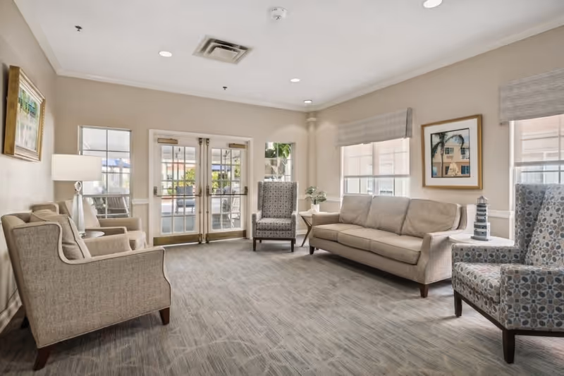 A bright communal sitting room with sofas, upholstered armchairs, side tables and double glass doors leading outside.