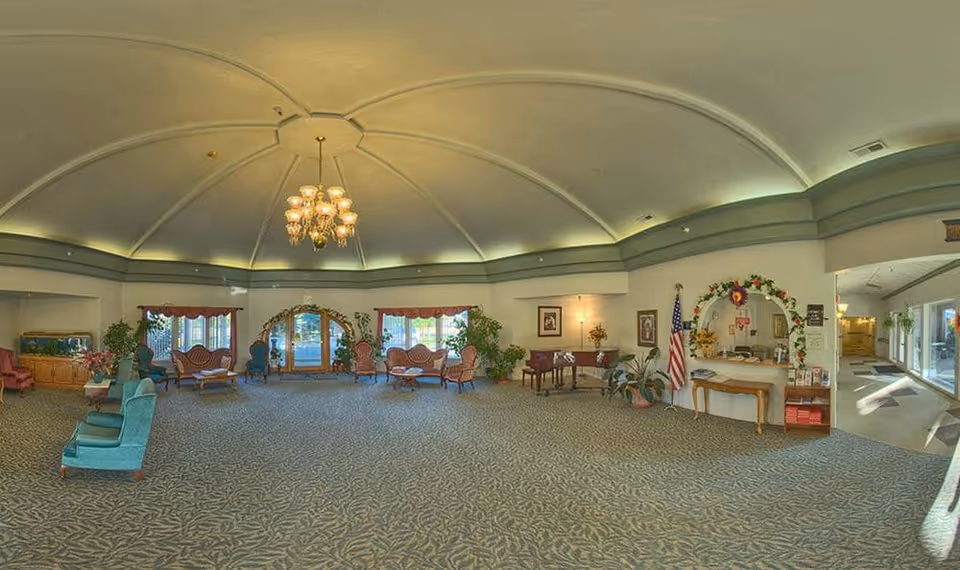 A spacious, well-lit assisted living facility common area with a high domed ceiling and chandelier. The room features multiple seating arrangements with vintage-style chairs and sofas, potted plants, framed pictures on the walls, an American flag, and a small reception desk decorated with flowers and brochures. Large windows with red valances allow natural light to enter, and there is a hallway leading to other parts of the facility.