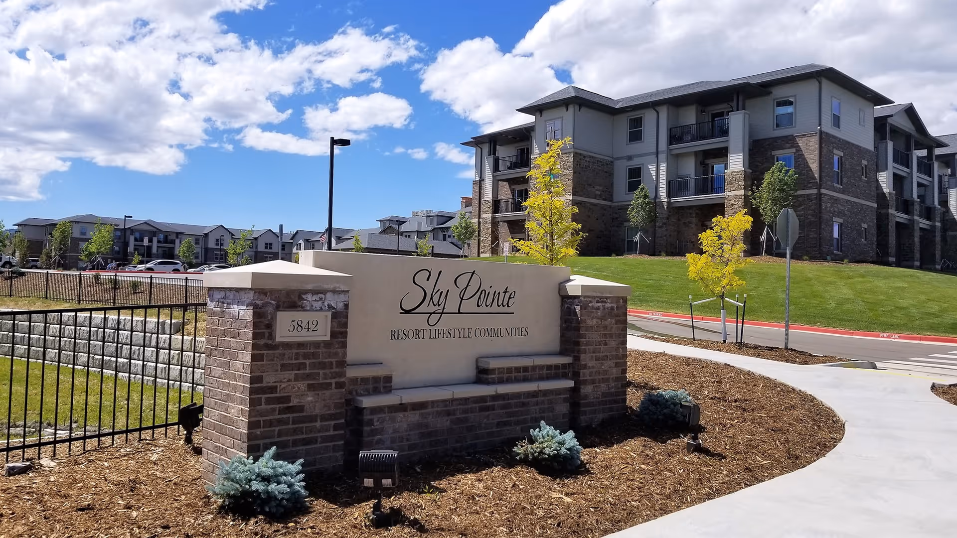 Entrance sign for Sky Pointe Resort Lifestyle Communities made of brick and stone with landscaping around it, with a multi-story residential building and a clear blue sky with clouds in the background.