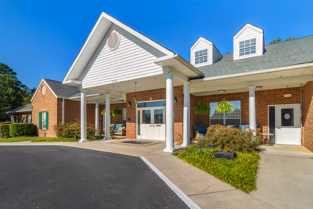 Front exterior view of a brick building with white columns supporting a covered entrance. The building has a green roof with dormer windows and hanging plants near the entrance. There is a paved driveway and sidewalk leading to the entrance.