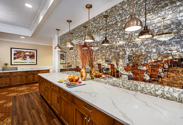 A well-lit dining room area with a long marble countertop island featuring wooden cabinets underneath. Above the island, there are six hanging pendant lights with bronze finishes. The back wall is covered with a reflective, textured mirror. On the countertop, there are decorative vases with red branches and a tray with fruit and snacks. In the background, tables and chairs are visible, set for dining.